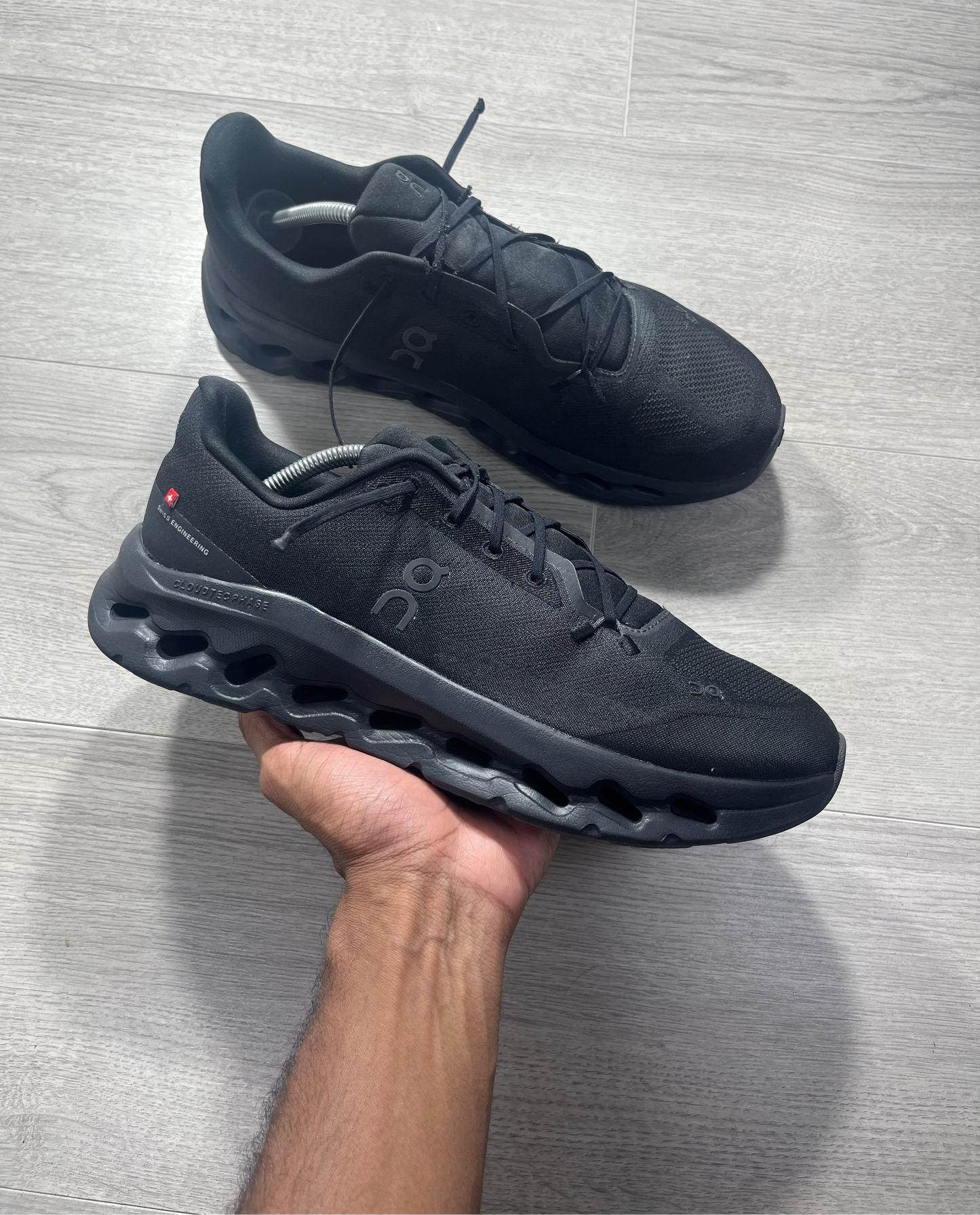 A pair of black On running shoes, with one held up by a hand and the other placed on a light gray wood floor. The On logo is visible on the side of the shoe.