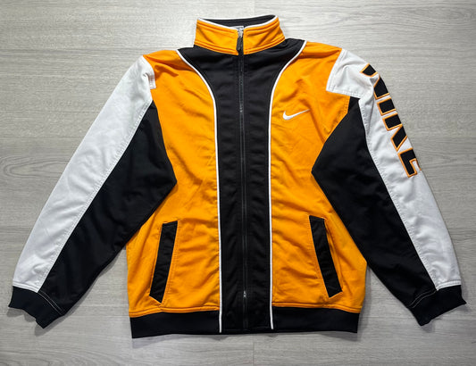 A Nike track jacket laid flat on a gray wood-textured surface. The jacket features a bold color-blocked design with a bright yellow-orange body, black center panel and cuffs, white sleeves, and black-and-orange striped detailing on the right sleeve.
